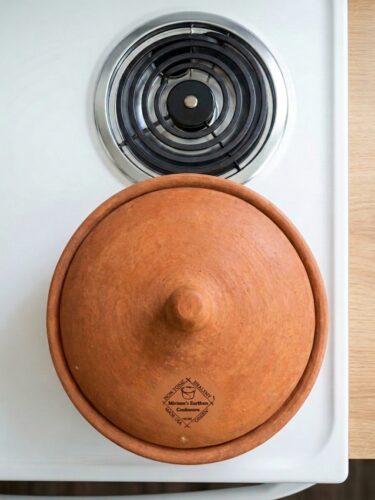Top-down view of a round Miriam's pure clay Dutch oven with a matching domed lid, centered on a white electric coil stovetop. The lid features a small knob handle and the Miriam's Earthen Cookware logo near the bottom. The warm, natural clay contrasts with the clean white stove and black burner, creating a simple, minimalist composition.