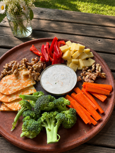 Pure clay griddle used as a serving platter on a wooden picnic table holding fresh broccoli, carrot sticks, red bell peppers, cubed and sliced cheese, walnuts, and a bowl of creamy dip, with a jar of daisies nearby.