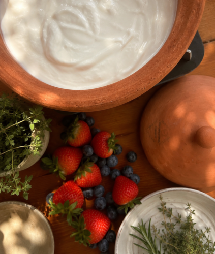 Creamy homemade yogurt in an unglazed pure clay pot on a wooden counter, with the clay lid, and a small pile of fresh strawberries, blueberries, and raspberries alongside.