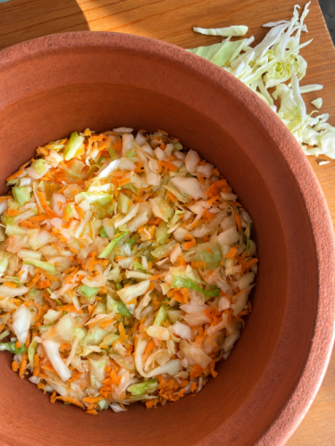 Freshly mixed shredded cabbage and grated carrot for sauerkraut in a terracotta clay pot, with a pile of sliced cabbage on a wooden cutting board beside it.