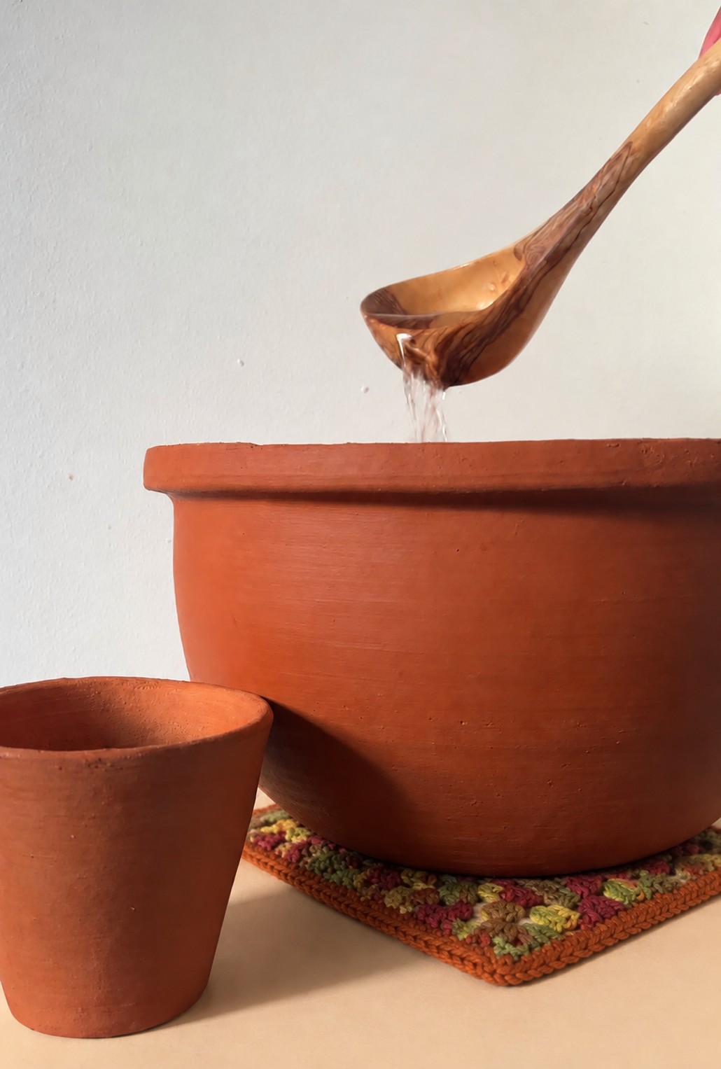 A pure clay water jar sitting on a hand-crocheted cotton coaster in warm autumn colors, with water being ladled in from an olive-wood spoon and a small clay cup beside it.